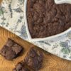 A heart-shaped dish filled with freshly baked brownies sits atop a colorful floral towel, with three brownies arranged beside it, showcasing their rich, fudgy texture.