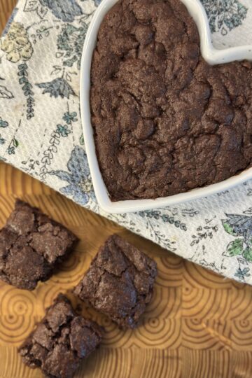 A heart-shaped dish filled with freshly baked brownies sits atop a colorful floral towel, with three brownies arranged beside it, showcasing their rich, fudgy texture.