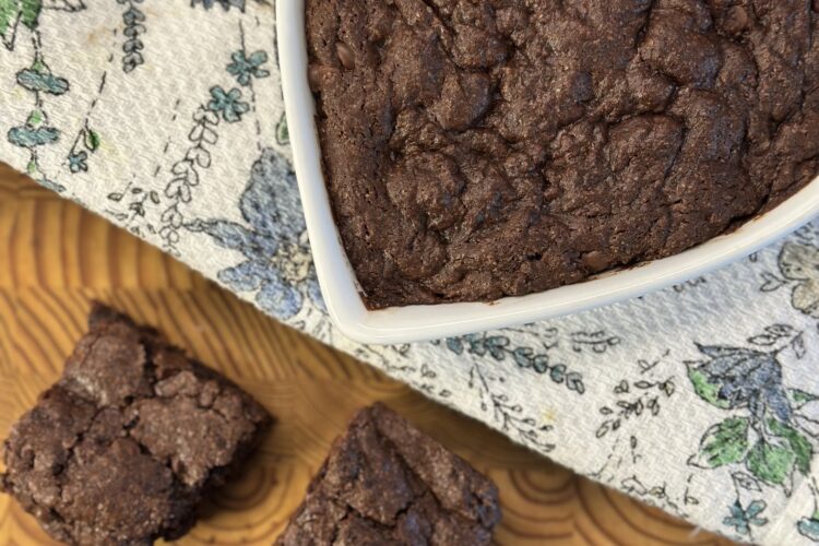 A heart-shaped dish filled with freshly baked brownies sits atop a colorful floral towel, with three brownies arranged beside it, showcasing their rich, fudgy texture.