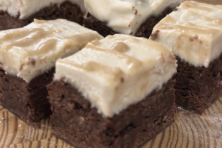 Vegan brownie cheesecake bars cut into squares and displayed on a rustic wooden cutting board, drizzled with a rich caramel sauce