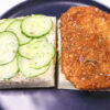 Open-face Vegan Cornflake "Chicken" Sandwich on a plate, showcasing crispy tofu with garlic butter sauce, creamy cashew spread, marinated cucumber, and red onion.