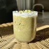 A bowl of vegan pistachio chia seed pudding on a wicker table, adorned with whipped cream, chopped pistachios, and white chocolate.