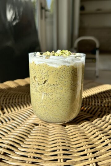 A bowl of vegan pistachio chia seed pudding on a wicker table, adorned with whipped cream, chopped pistachios, and white chocolate.